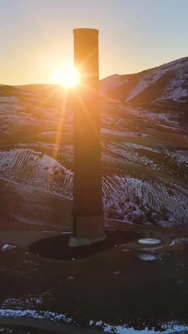The low sun shines past a tall brick smelter stack above snowy ground.