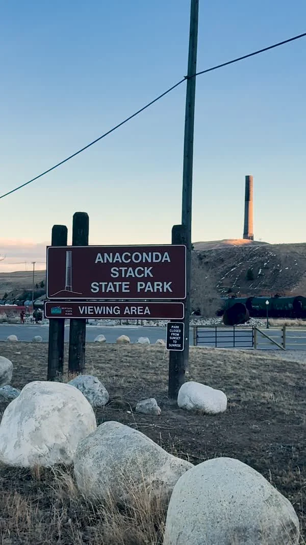 An Anaconda Stack State Park viewing-area sign stands below the distant stack.