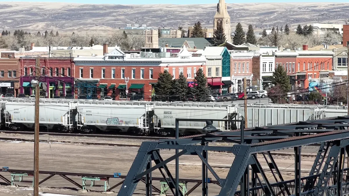 A pedestrian walks a dog across a steel footbridge above railway tracks, with Laramie storefronts and hills beyond.