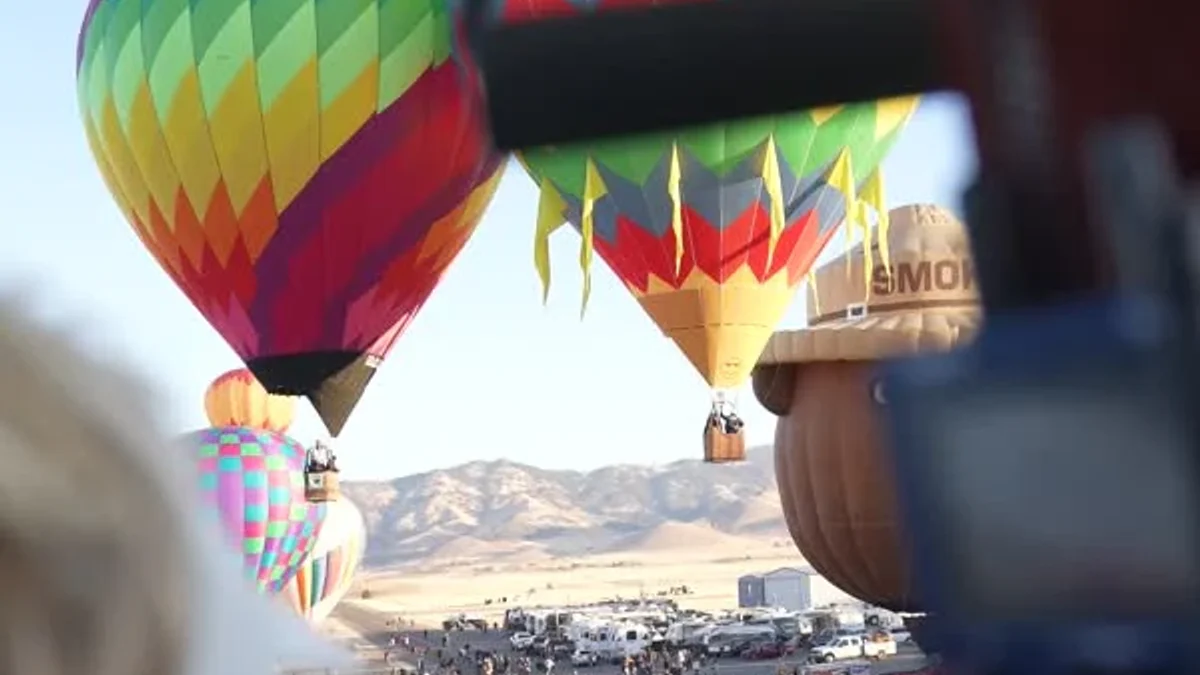 Colorful hot-air balloons lift above a festival crowd, seen past a basket and spectators.
