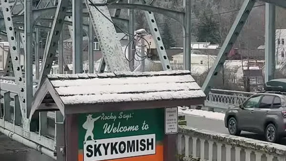 Snow-dusted Skykomish storefronts and its welcome sign sit beneath forested mountain slopes.