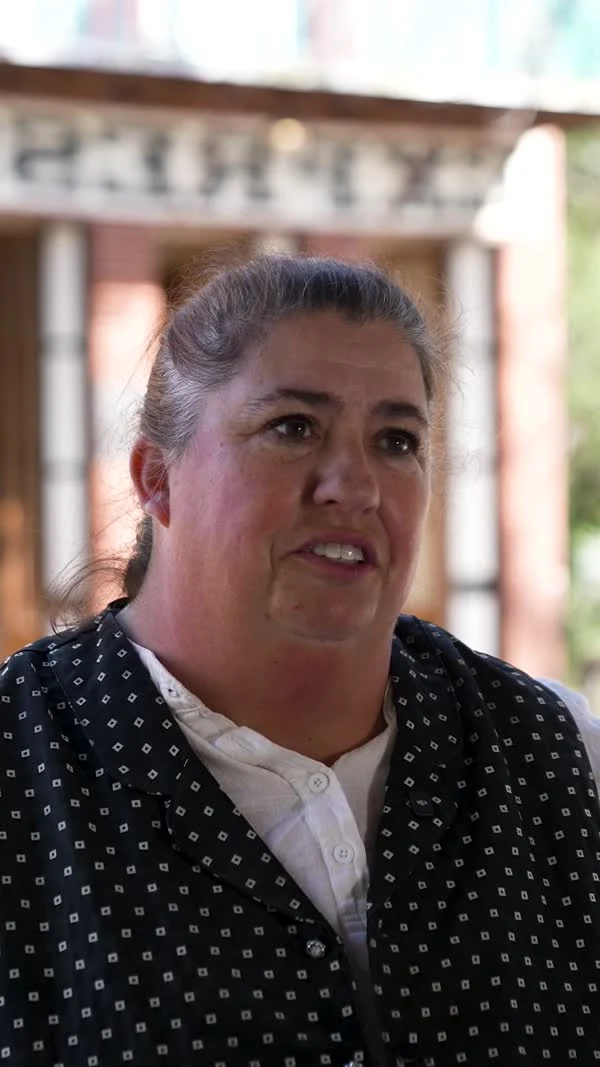 A participant in a dark vest speaks outside a wooden building.