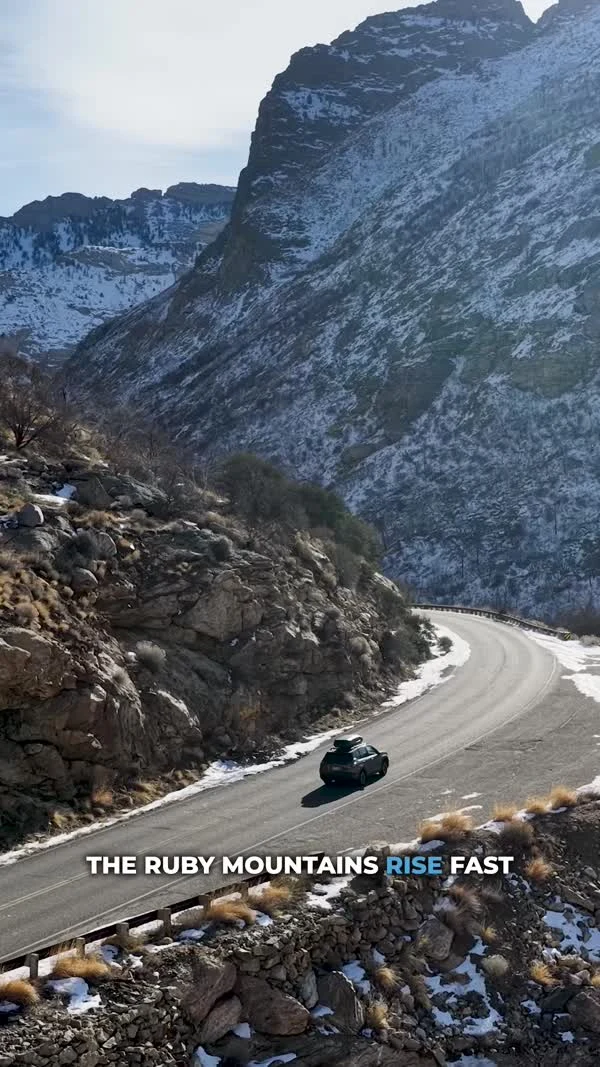 A car follows a winding road between steep, snow-dusted mountain slopes.