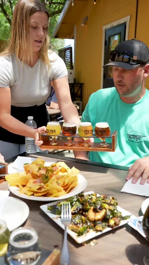 A server sets a beer tasting flight beside a seated diner and plates of food.