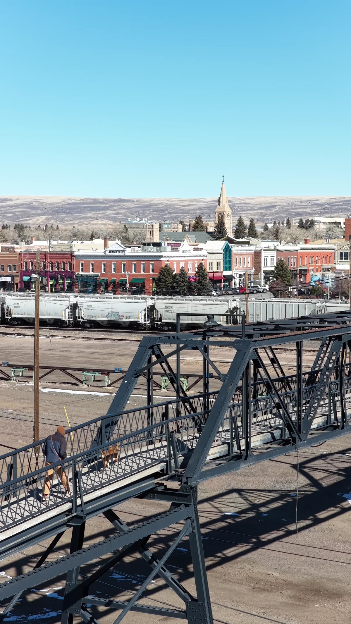 A pedestrian walks a dog across a steel footbridge above railway tracks, with Laramie storefronts and hills beyond.