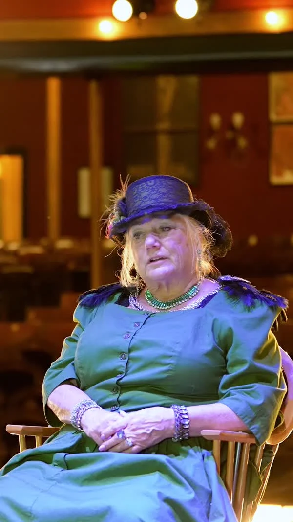 A participant wearing a green dress and blue hat sits on a stage under theater lights.