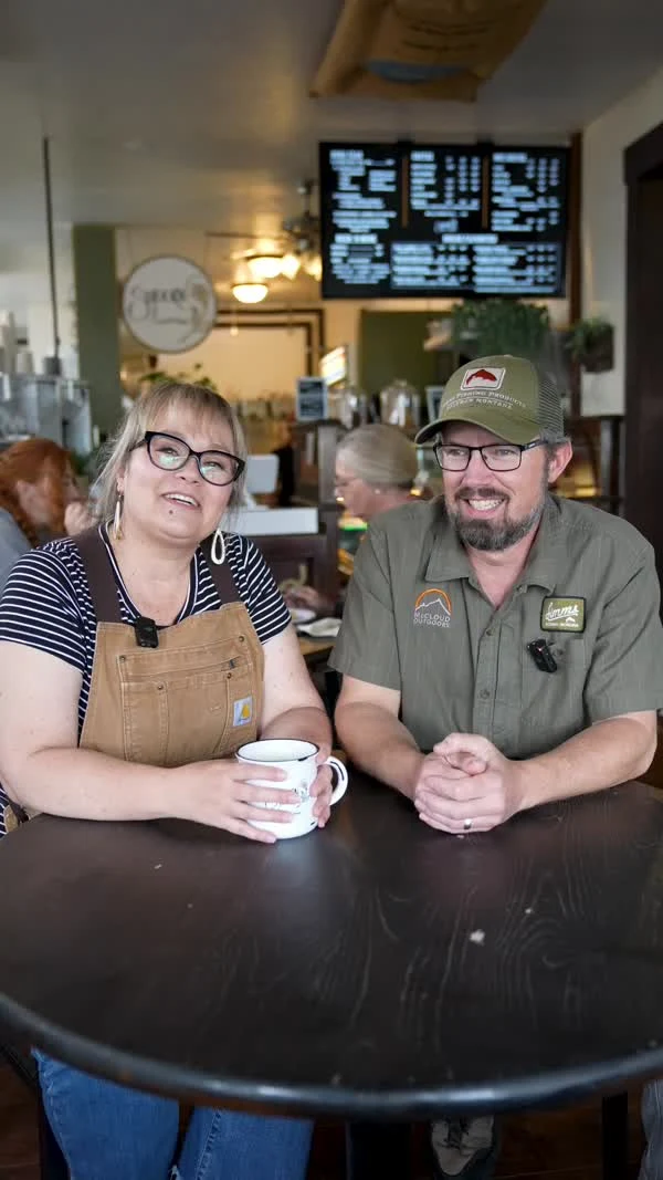 Two participants sit together at a coffee-shop table, smiling toward the camera.
