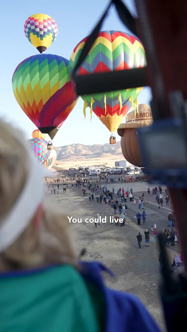 Colorful hot-air balloons lift above a festival crowd, seen past a basket and spectators.