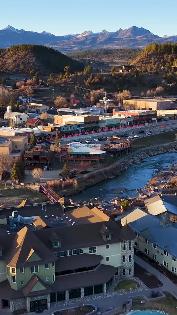 An aerial view follows the river past colorful buildings toward distant mountains.
