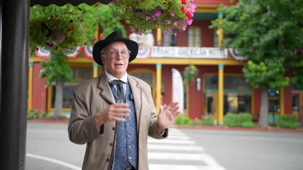 A presenter in a dark hat and tan jacket speaks on a tree-lined downtown street.